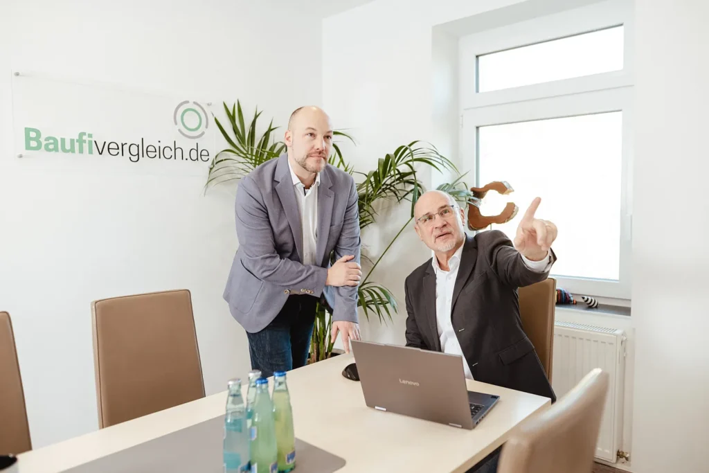 Die Geschäftsführer und Immobilienberater Salomon Davin (links) und Holger Böttger (rechts) von Baufivergleich.de im Beratungsgespräch am Laptop im Büro in Kassel, im Hintergrund das Firmenlogo.