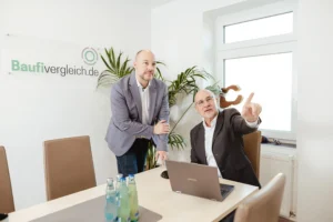 Die Geschäftsführer und Immobilienberater Salomon Davin (links) und Holger Böttger (rechts) von Baufivergleich.de im Beratungsgespräch am Laptop im Büro in Kassel, im Hintergrund das Firmenlogo.