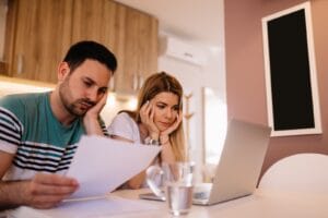 Paar sitzt zu Hause am Tisch und prüft die Unterlagen zum Bausparvertrag, im Hintergrund ein Laptop