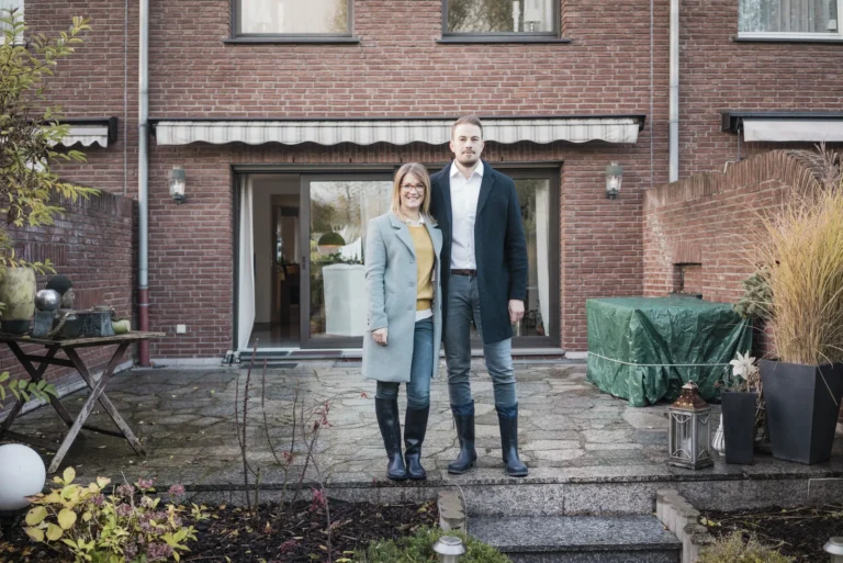 Junges Paar mit Unterlagen vor einem Einfamilienhaus prüft die teuersten Fehler beim Hauskauf